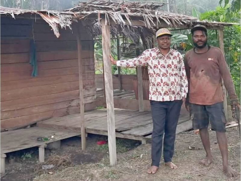 Kepala Distrik Tomu, Syamsul Inai (berbaju batik), berfoto bersama warga dengan latar belakang rumah pondok tanpa penerangan.
Foto: Dokumen Kepala Distrik Tomu.  